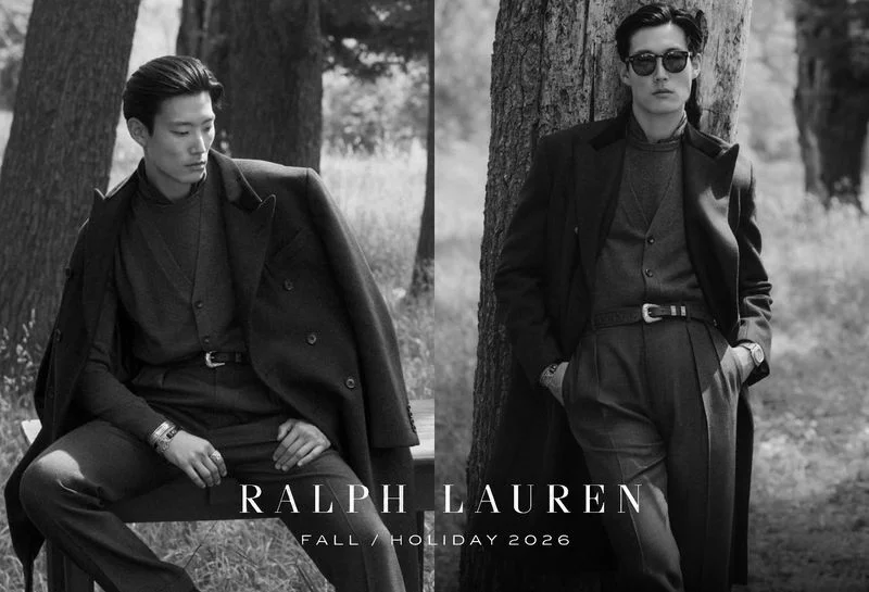 Black and white fashion campaign image featuring two male models in tailored coats and trousers outdoors near trees