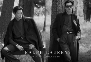 Black and white fashion campaign image featuring two male models in tailored coats and trousers outdoors near trees