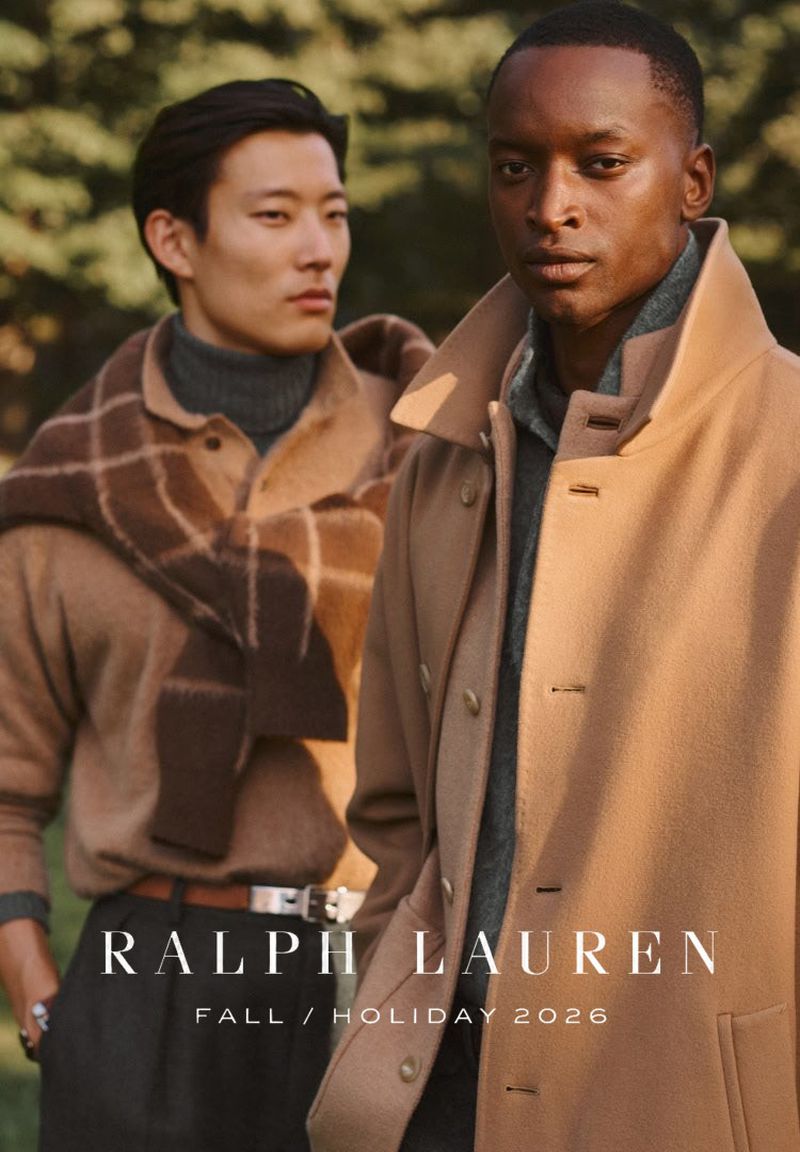 Two male models in camel coats and sweaters standing outdoors against a natural pine tree background.