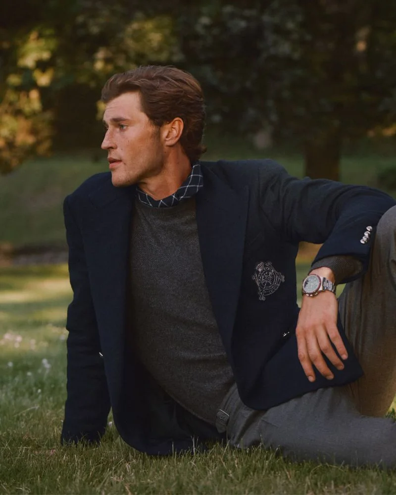 Man wearing a dark blazer, grey sweater, and patterned shirt collar seated outdoors on a lawn for Ralph Lauren