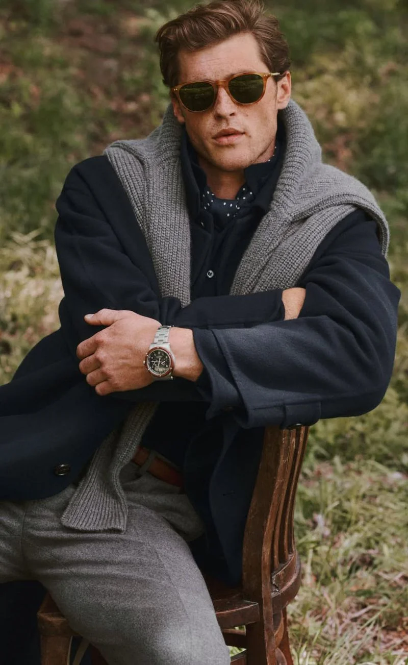 Model wearing sunglasses and a navy coat with a grey knit sweater draped over shoulders seated outdoors