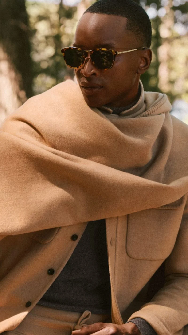 Model wearing sunglasses and a camel jacket draped in a scarf outdoors