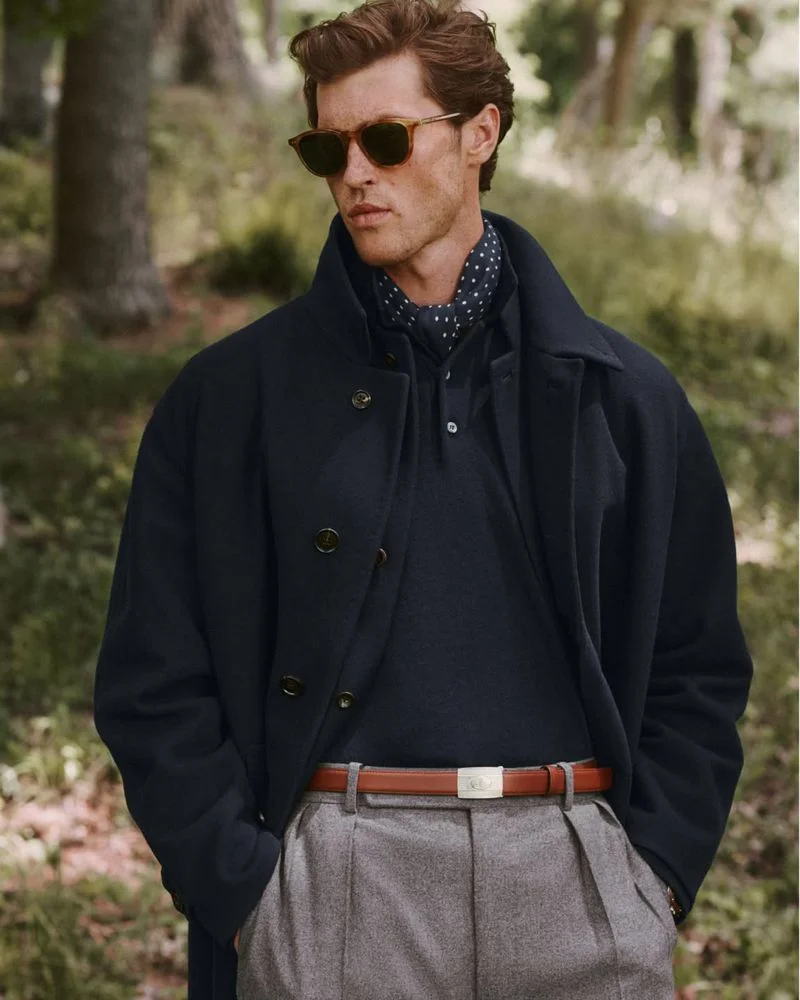 Model in sunglasses and dark coat with a patterned scarf standing outdoors in front of trees for Ralph Lauren