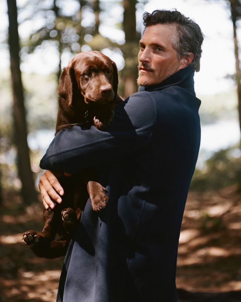 Man wearing a navy coat holding a brown labrador puppy outdoors in a wooded forest setting.