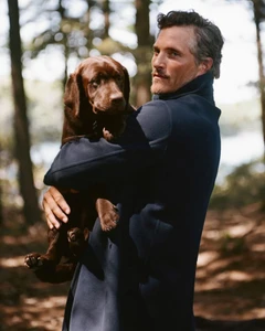 Man wearing a navy coat holding a brown labrador puppy outdoors in a wooded forest setting.