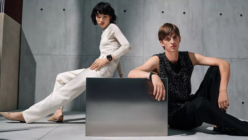 Two models posing against a grey concrete wall featuring a metallic block in the foreground for Hermès Apple Watch 2026 campaign.