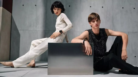 Two models posing against a grey concrete wall featuring a metallic block in the foreground for Hermès Apple Watch 2026 campaign.
