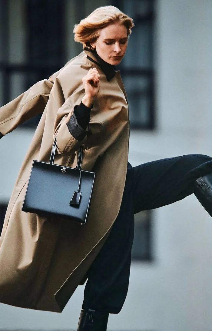Model in a tan coat carrying a black handbag while moving dynamically outdoors
