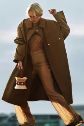 Model in a long brown coat holding a brush-shaped bag against a clear sky background