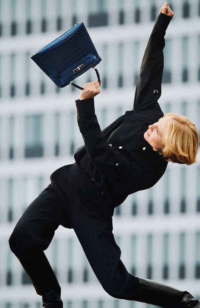 Model in black clothing holding up a blue handbag against an urban building facade