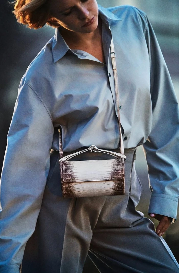 Model wearing a light blue button-down shirt and grey trousers with a textured brown and white crossbody bag