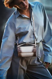 Model wearing a light blue button-down shirt and grey trousers with a textured brown and white crossbody bag