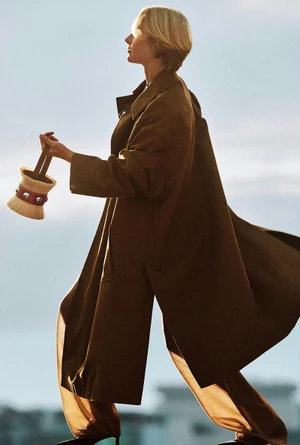 Model in profile wearing a long brown coat walking outdoors and holding a small bucket-shaped bag against a clear sky background
