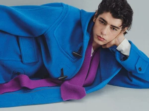 Model in vibrant blue duffel coat with purple scarf resting head on hand against a pale grey studio background.