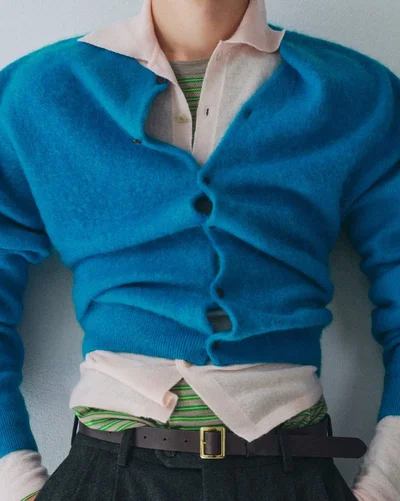 Close-up of a person wearing a bright blue cardigan over layered light pink and striped shirts with dark trousers.
