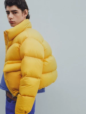 Close-up portrait of a male model wearing a bright yellow puffer jacket against a solid light blue background.
