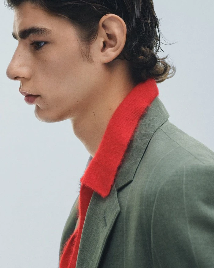 Profile portrait of a male model wearing an olive green blazer and a vibrant red collared shirt against a white backdrop.