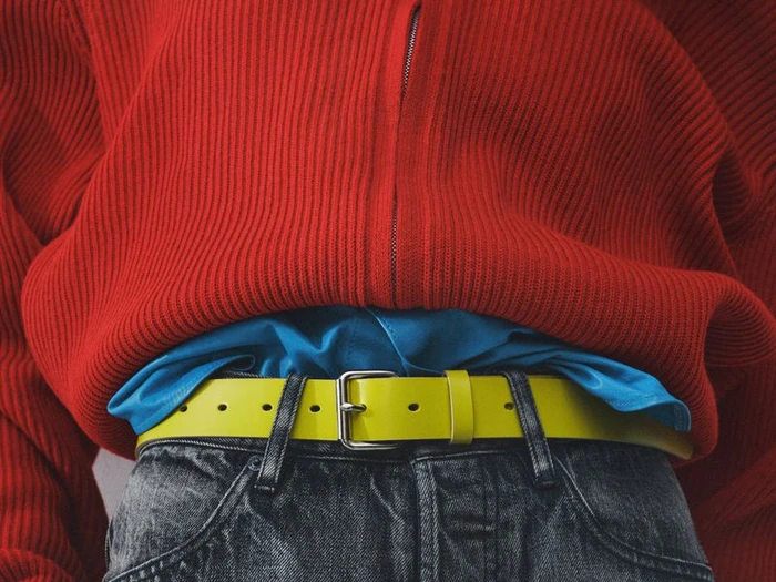 Close-up of a person wearing a bright red ribbed zip-up sweater over a blue shirt, dark jeans, and a yellow belt.