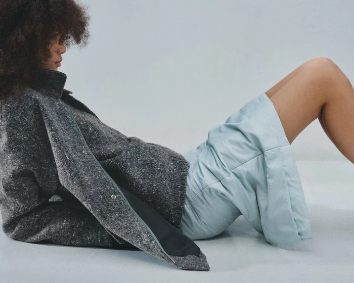 Model with dark curly hair wearing a grey textured jacket and pale blue shorts against a light studio backdrop
