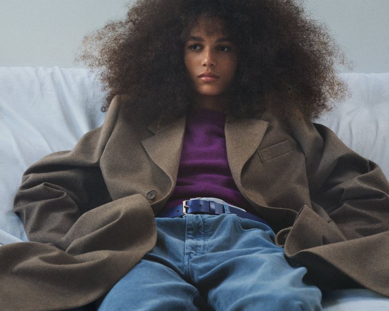 Model with voluminous curly hair wearing a brown oversized coat and purple sweater against a light background