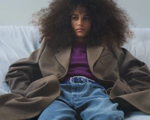 Model with voluminous curly hair wearing a brown oversized coat and purple sweater against a light background