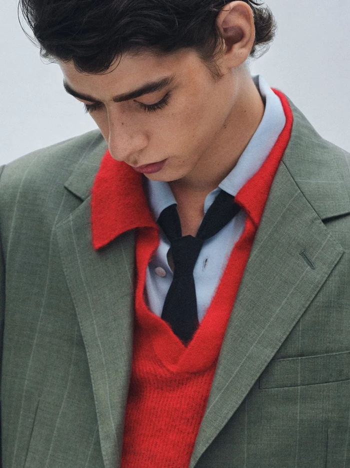 Close-up portrait of a young man wearing a green blazer over a red sweater, light blue shirt, and dark tie.