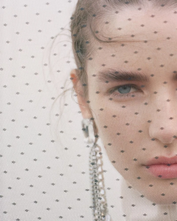 Close-up portrait of a woman looking through a dotted veil wearing dangling rhinestone earrings for H&M Studio AW 2026