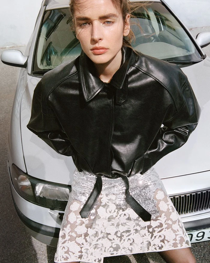 Model in black leather jacket and sheer patterned skirt leaning against a car for H and M Studio AW 2026 campaign.