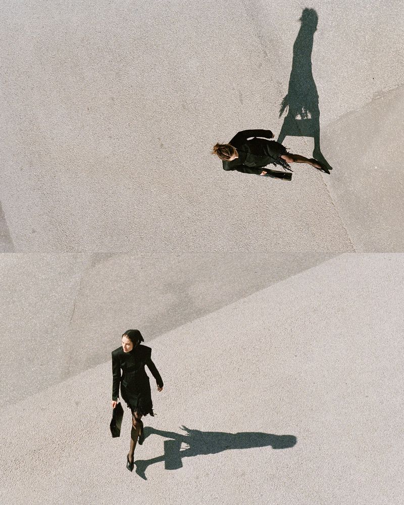 High-angle view of a model in a black dress carrying a handbag walking across a textured concrete plaza, casting a long shadow.