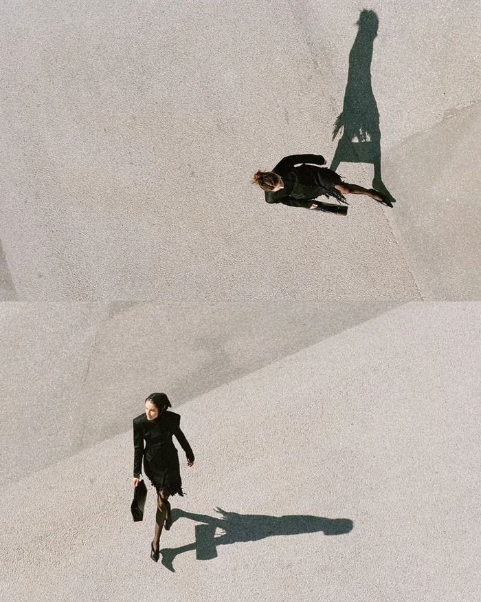 High-angle view of a model in a black dress carrying a handbag walking across a textured concrete plaza, casting a long shadow.