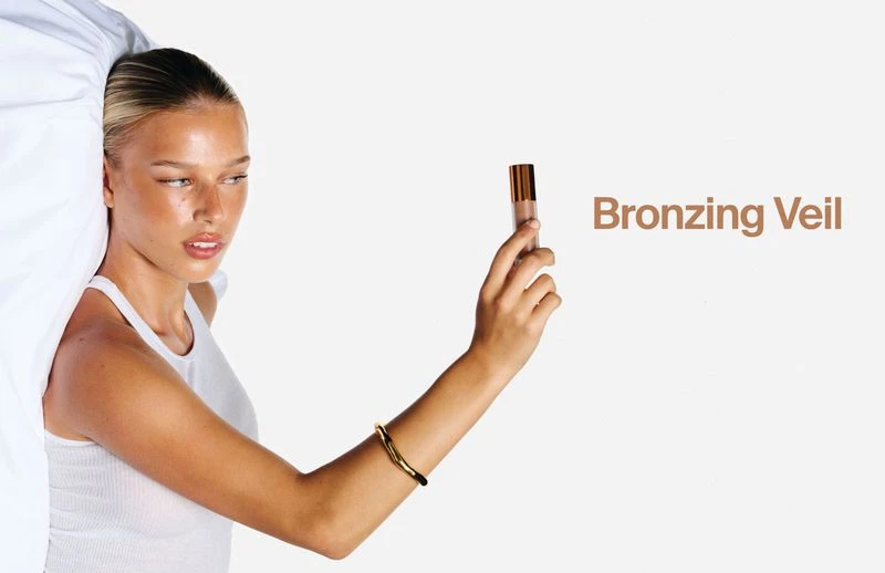 Model holding a small cosmetic bottle against a white background with visible typography for Haus Labs Bronzing Veil.