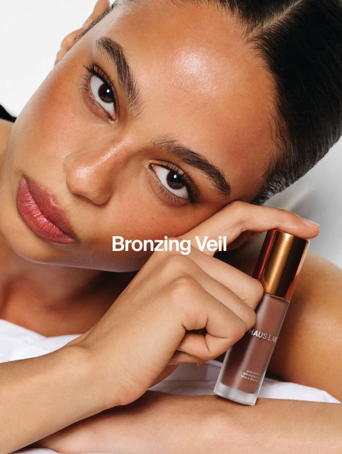 Close-up portrait of a model holding a Haus Labs bronzer product bottle against a seamless white background.