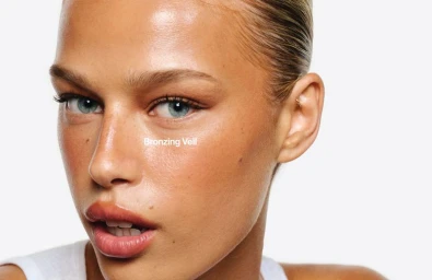 Close-up portrait of a model with glowing bronzed skin and blue eyes against a white background for Haus Labs Bronzing Veil campaign.