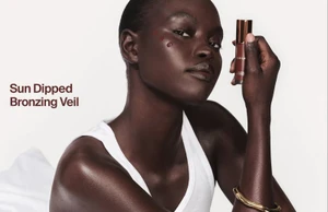 Model wearing a white tank top holding a bronzing product tube against a white background for Haus Labs