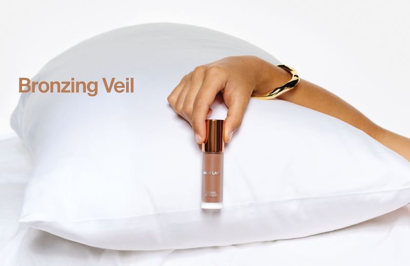 Hand holding a bronze cosmetic bottle against a white satin pillow for Haus Labs Bronzing Veil campaign