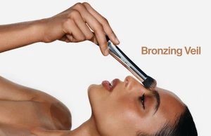 Close-up profile of a model holding a makeup brush near her nose against a white background with brown text