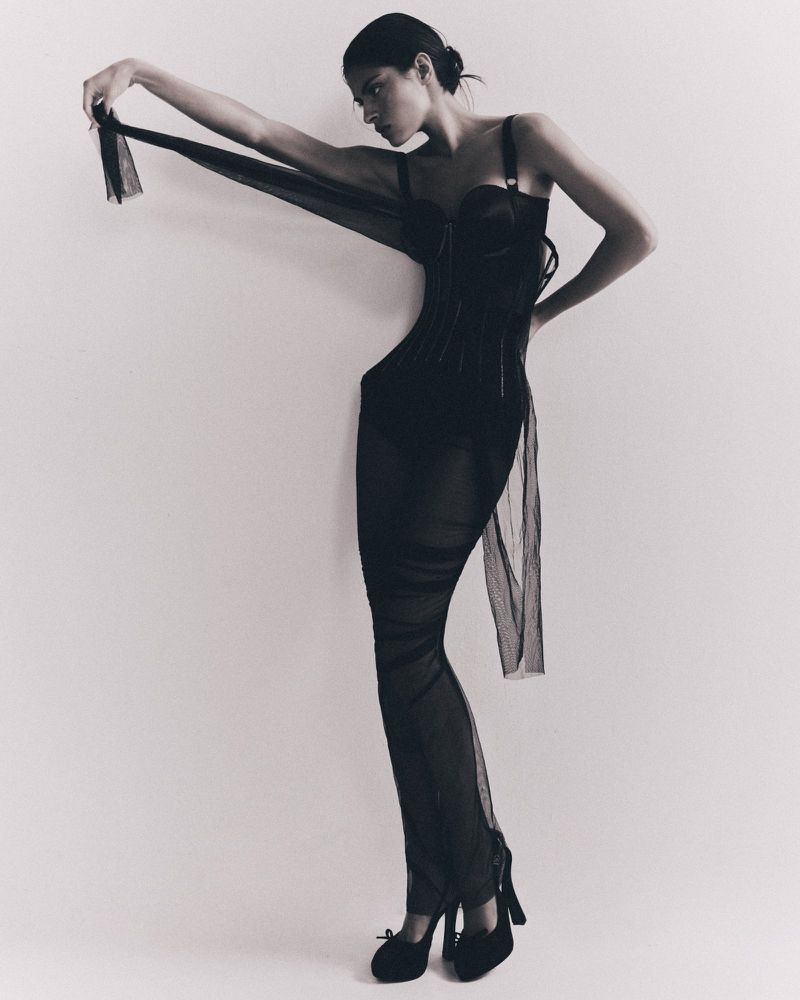 An adult female fashion model poses in a full-length shot, wearing a black semi-sheer dress and heels against a white background.