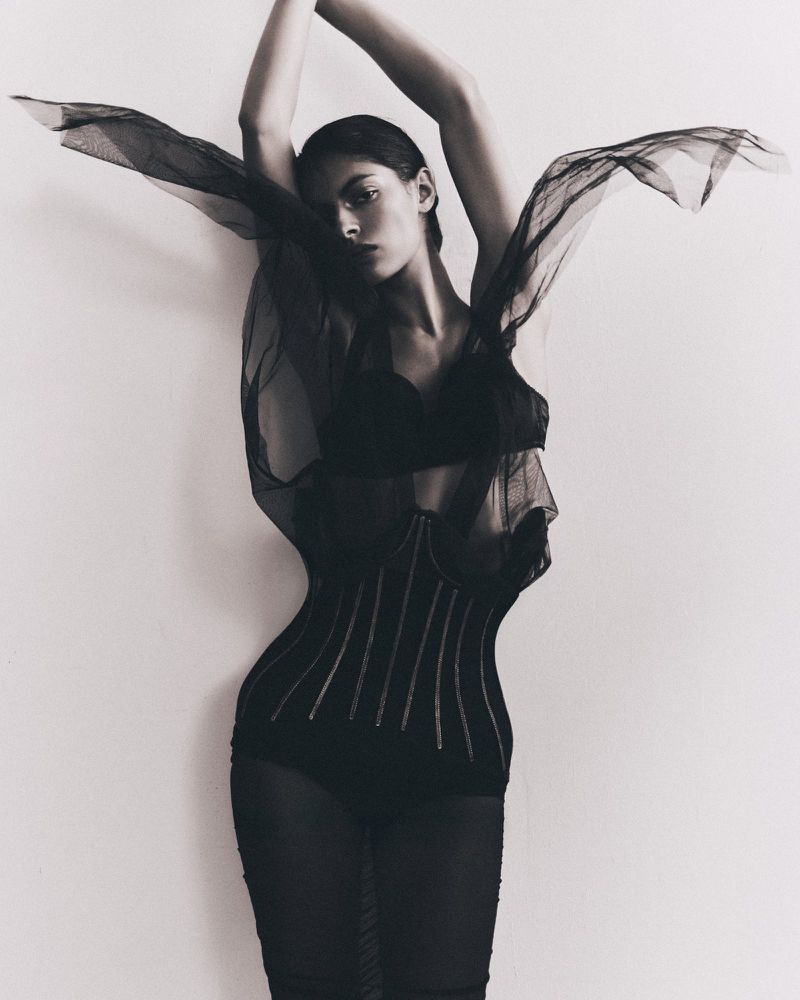 A woman strikes a dramatic pose indoors, wearing a black, semi-sheer garment, against a light wall, creating a striking fashion portrait.