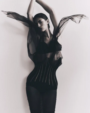 A woman strikes a dramatic pose indoors, wearing a black, semi-sheer garment, against a light wall, creating a striking fashion portrait.