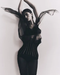 Model in black sheer dress with arms raised against a plain white studio background