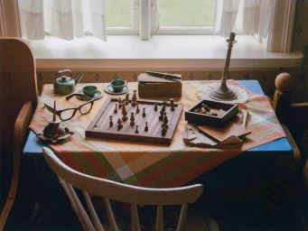 Wooden chess set on a small table with tea cups, glasses, and a candlestick in front of a window