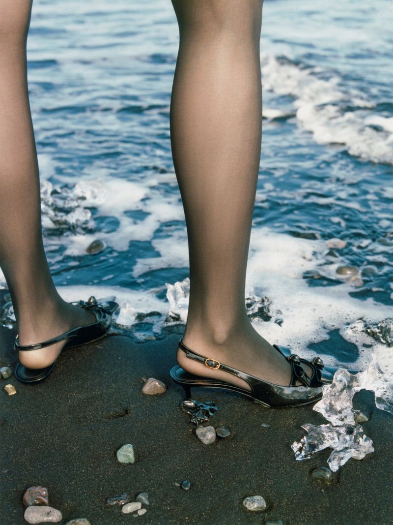 Close-up of legs in black stockings and black slingback heels standing on a dark sandy beach with ocean waves.