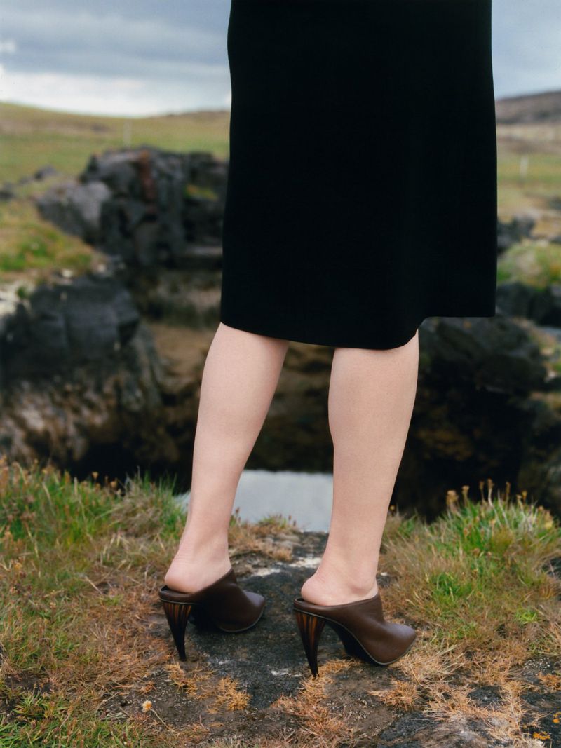 Model standing outdoors wearing a black skirt and brown heeled mules against a rocky landscape background