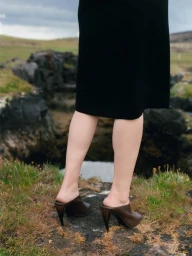 Model standing outdoors wearing a black skirt and brown heeled mules against a rocky landscape background