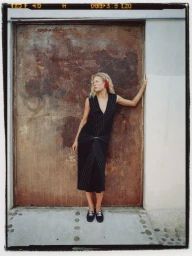 Model wearing a dark sleeveless dress and loafers leaning against a weathered rust door for InStyle Mexico
