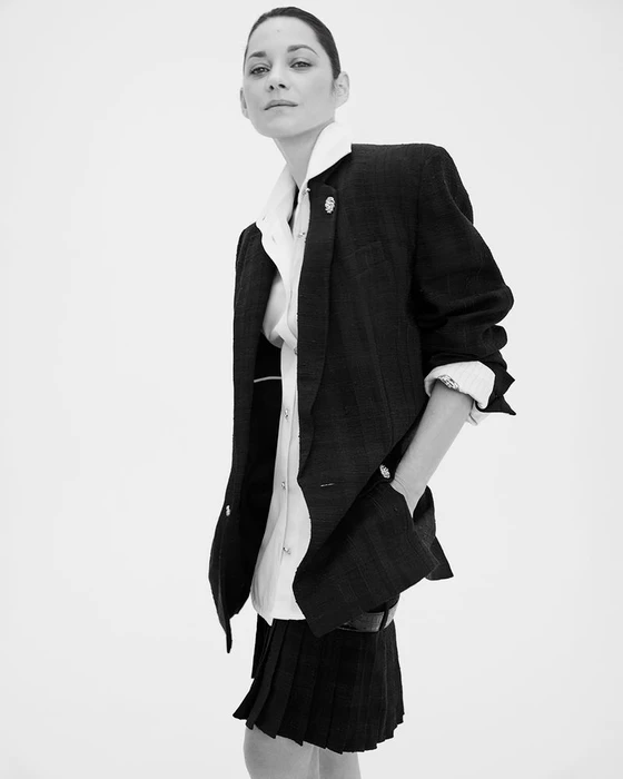 Marion Cotillard wearing a dark blazer over a white shirt and pleated skirt against a plain white background