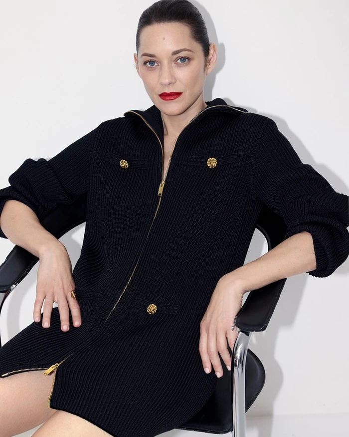 Marion Cotillard sitting in a black chair wearing a black knitted zipped dress against a white background.