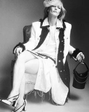 Vanessa Paradis seated in an armchair wearing a white tweed coat with black trim, holding a black handbag.