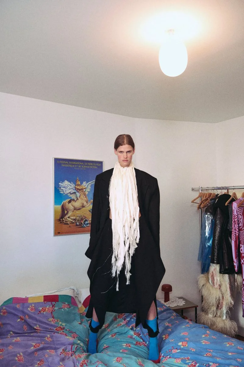 Model standing on a patterned blue bed wearing a black coat and a long white scarf in a bedroom setting