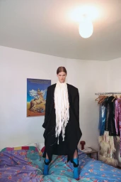 Model standing on a patterned blue bed wearing a black coat and a long white scarf in a bedroom setting